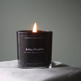 A Predominantly Black scented candle named "Solitude," with a gently flickering flame, casting a soft light on a peaceful setting.