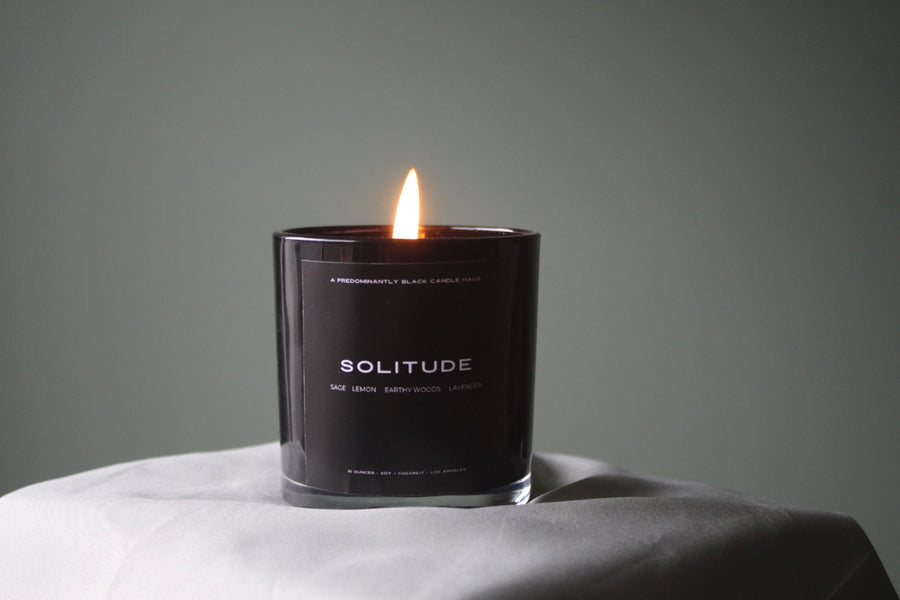 A Predominantly Black scented candle named "Solitude," with a gently flickering flame, casting a soft light on a peaceful setting.