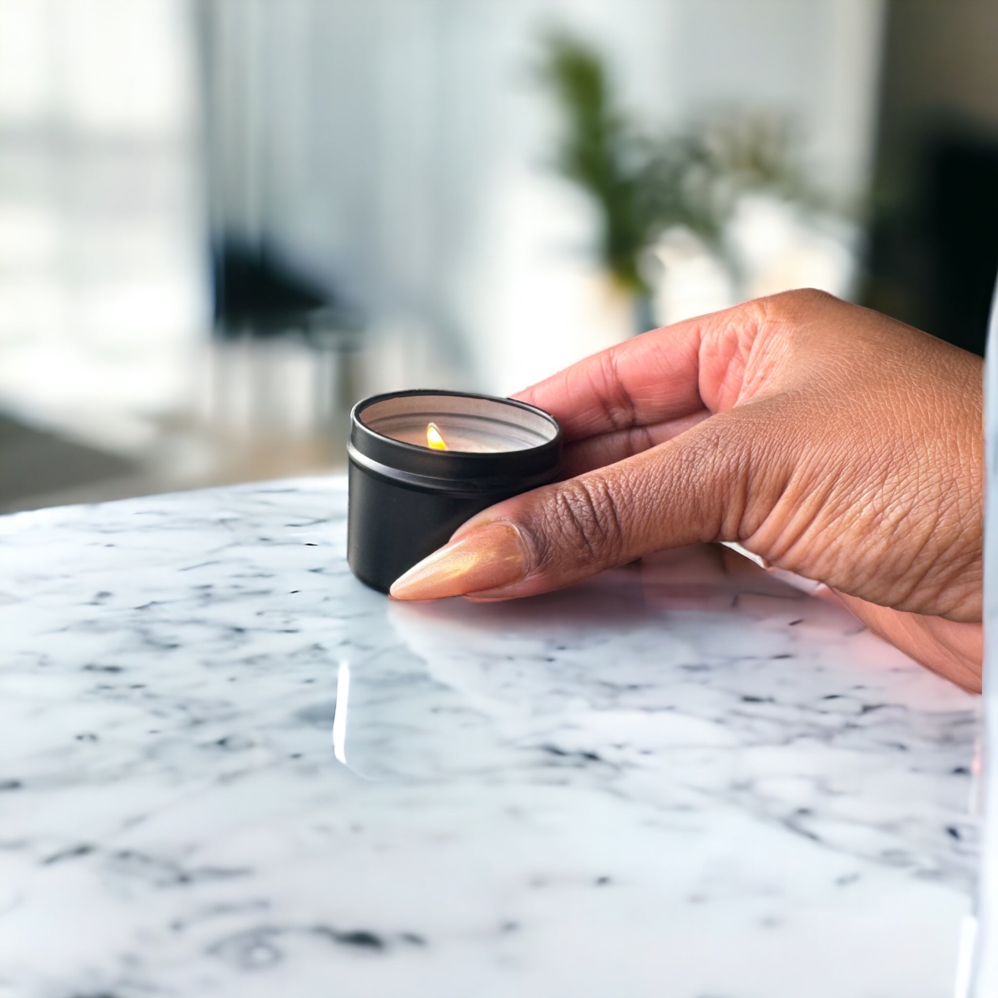 A person's hand with manicured nails gently holding a Predominantly Black A TINY TIN CANDLE - 06483 on a marble surface, conveying a sense of warmth and tranquility.