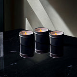 Three Predominantly Black A TINY TIN CANDLES - 06483, including a pine scented candle, on a dark surface with a striking interplay of light and shadow.