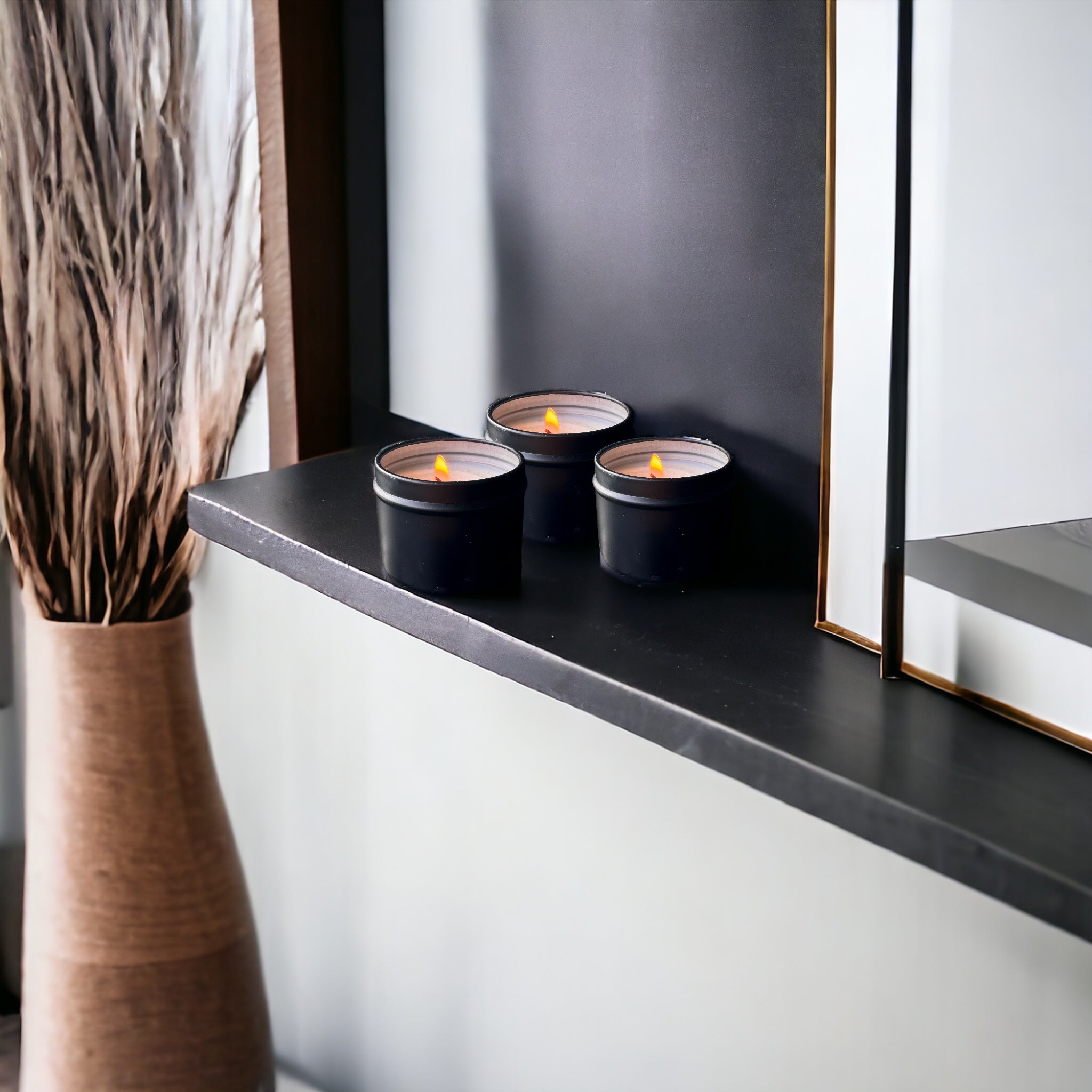 Three lit A TINY TIN CANDLES - MADNESS CANDLES, including a tropical paradise candle, on a modern shelf, creating a tranquil and cozy ambiance next to a decorative mirror and vase with dried twigs.