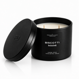 A matte black jar candle with its lid leaning against it on a white studio background. The front label reads 'BISCOTTI MAMI' and 'ALMOND, PECAN, COCO BUTTER, VANILLA', and the lid has an embossed 'PB' logo.