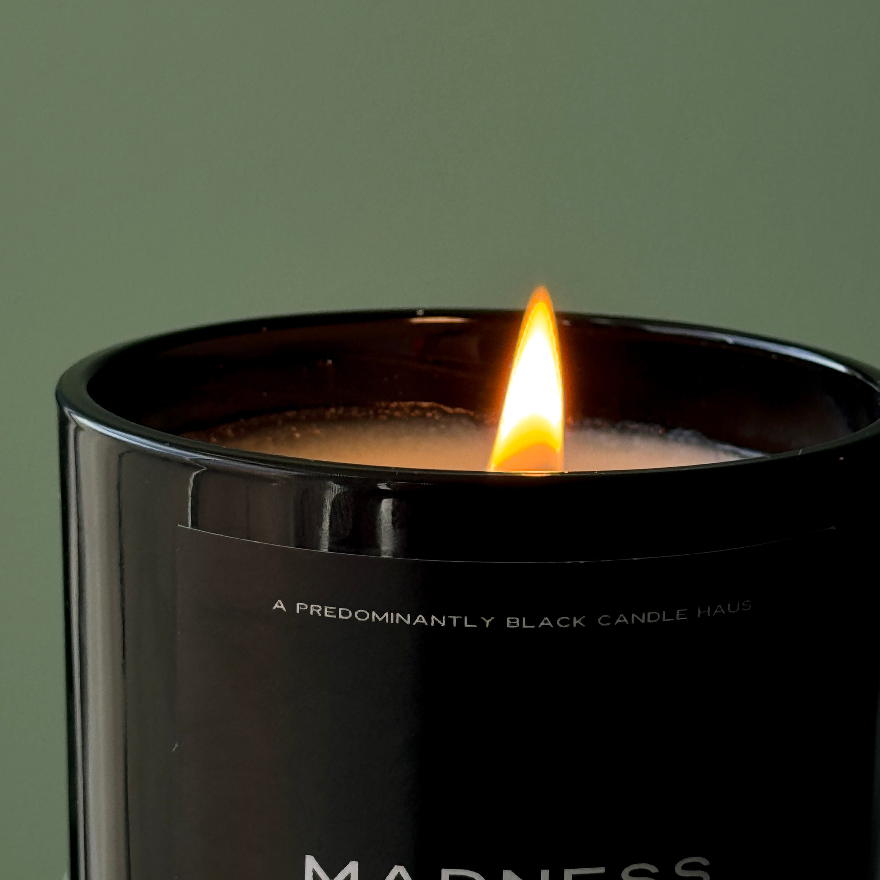 A glowing candle flame atop a Predominantly Black A MADNESS CANDLE with a description of its various tropical paradise scented notes.