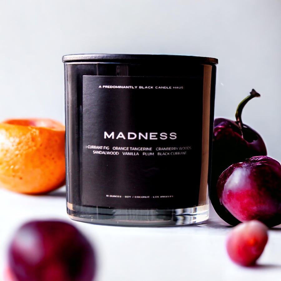 A sleek black luxury Predominantly Black candle with "madness" text   and a description of aromatic notes  positioned infront of  citrusy fruits like plums and oranges.